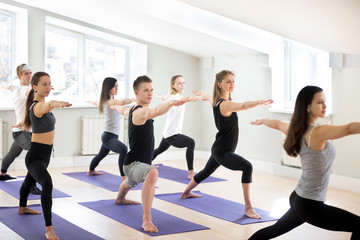 Fototapeta na wymiar Group of sporty people practicing yoga lesson with instructor, doing Warrior Two pose, Virabhadrasana 2 exercise, indoor session close up, students working out in sport studio. Wellbeing lifestyle