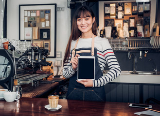 Asian woman barista wear jean apron holding blank screen tablet to customer at bar counter with smile emotion,Cafe restaurant service concept,Owner small business.mock up device for display text.