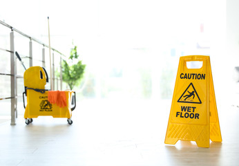 Phrase "CAUTION WET FLOOR" on safety sign and mop bucket with cleaning supplies, indoors © New Africa