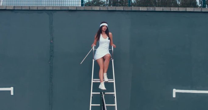 Portrait of young Caucasian teen model wearing fashionable tennis dress, posing on tennis judge umpire chair, summer sunny day outdoors. Fashion portrait shoot