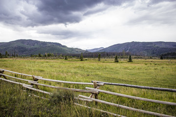 Fototapeta na wymiar Wood Fence Runs along a Meadow with the Rocky Mountains and Grey Clouds in the Distance, Colorado, USA