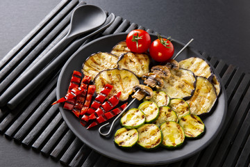 Grilled vegetables: eggplant, zucchini, pepper. Skewer with mushrooms. On a black plate and a black background.