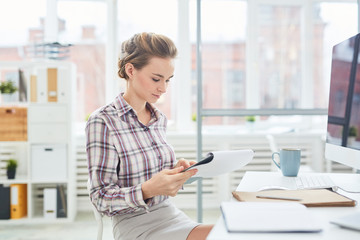 Obraz premium Young pretty Caucasian woman sitting at office desk and reading important paper on clipboard