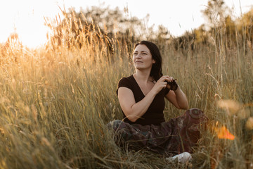 A portrait of a happy woman sitting in grass and doing her hair. A young woman enjoying plaiting hair in the nature at sunset.