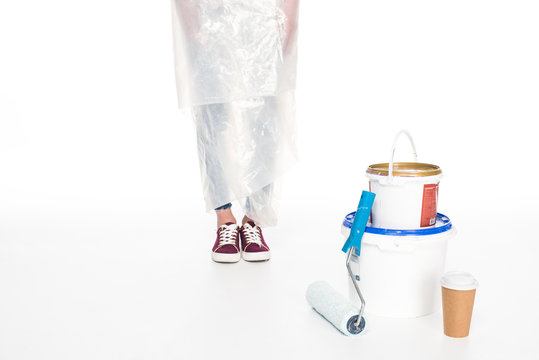 cropped image of woman in polyethylene cover standing near paint tins, paint roller and paper cup of coffee isolated on white background