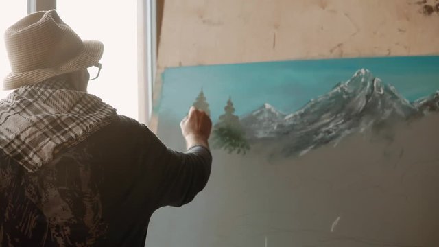 Inspired old painter is drawing spruces on a picture with mountains landscape. He is standing back to camera, and using artistic brush