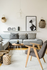 Grey armchair next to wooden table in bright interior with poster and cushions on sofa. Real photo