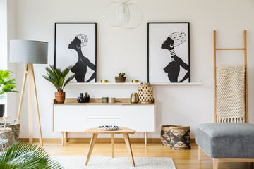Black and white african posters above cabinet in living room interior with lamp and table. Real...