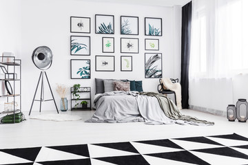 Real photo of a black and white bedroom with a bed standing between a lamp and a chair and next to a wall with plants paintings