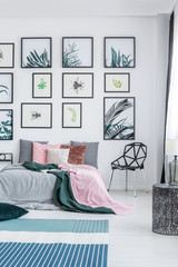 Real photo of a wide bed standing next to a black chair in a bedroom interior with plants posters on a wall and rugs on a floor