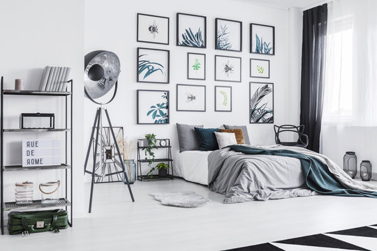 Real photo of a cozy bed standing next to a black lamp in a monochromatic bedroom with shelves with ornaments and posters on a wall