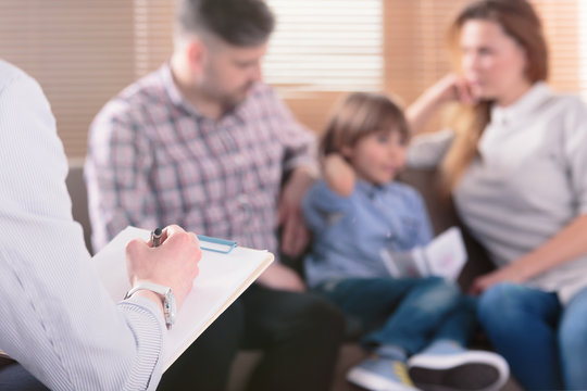 Hand of a professional family psychotherapist writing notes in front of a couple with a child in a blurred background during a consultation