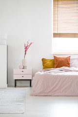 Real photo of a feminine bedroom interior with pink sheets on a bed standing near the window and wooden bedside cabinet with flowers