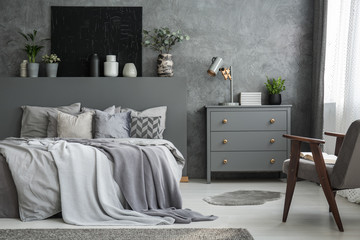 Grey cabinet next to bed in bedroom interior with armchair and black poster on the wall. Real photo