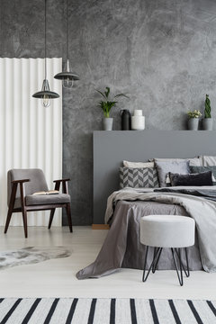 Striped carpet in grey bedroom interior with armchair next to bed against concrete wall. Real photo
