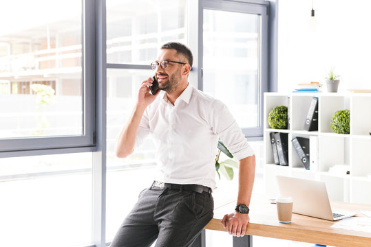 Image of happy smiling man in formal wear working and speaking on black smartphone, while standing near big window in office