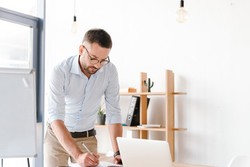 Photo of businessman 30s in white shirt writing down information at notebook from laptop, while working in bright office room