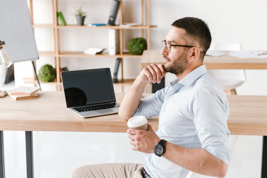 Profile image of diligent office man 30s in white shirt sitting at table and drinking takeaway coffee, while working on laptop in business centre