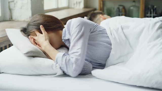 Sad woman is pajamas is lying in bed and crying after fight with her husband who is lying beside her with his back to her. Difficulties in married life concept.