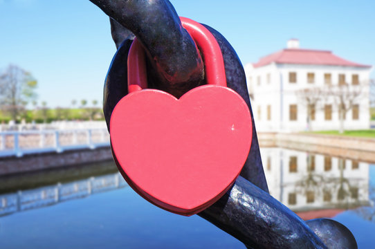 A heart shaped lock connected to chain, old house near the water defocused in the background.   