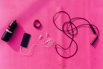 View from top on mobile phone with earphones, jump rope and bottle of water against the pink background. Sport concept.