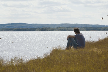 Fototapeta premium Young sad man sitting on the hill looks into the distance swallows flying over the river