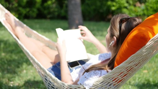 pretty young woman relaxing and reading a book in a hammock outdoor in the garden during a sunny summer day