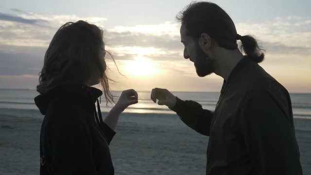 Attractive young couple sitting on the beach watching the sunset and making a heart shape with their hands in slow motion