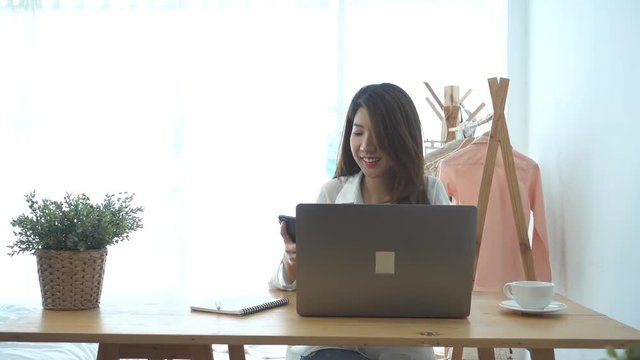 Slow motion - Beautiful young smiling asian woman working on laptop while sitting in a living room at home. Asian business woman using phone for work in her home office. Enjoying time at home.
