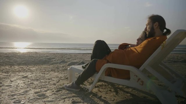 Affectionate young couple lying on a lounge chair on the beach watching the sunset while being covered with a blanket