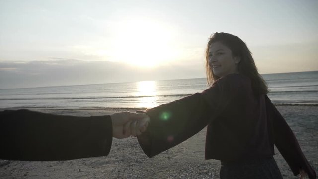 Young couple holding hands woman leading boyfriend walking towards sunset on empty beach POV travel concept in slow motion