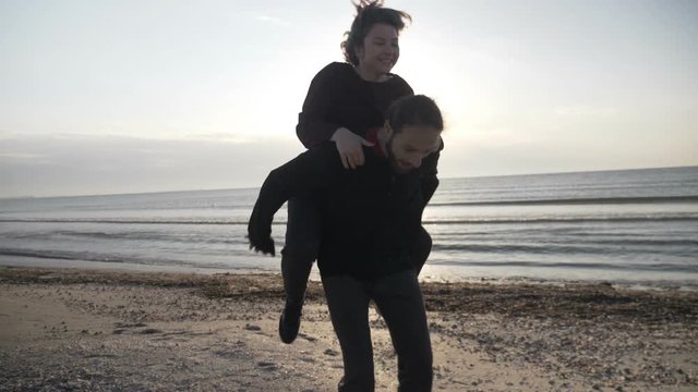 Young happy just married couple having fun on the beach playing piggy back during their honeymoon in slow motion