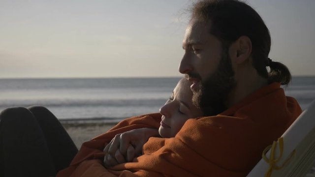 Close up of a young affectionate couple lying embraced on the lounge watching sunrise on the beach early in the morning covered with blanket in slow motion