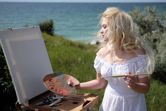 Beautiful girl draws a picture in the park using a palette with paints. Easel and canvas with a picture.