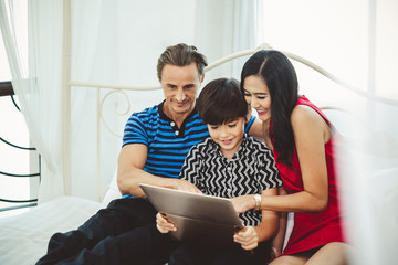 Mother, father and son, cute boy playing game with laptop on white bed, happy family and lifestyle concept