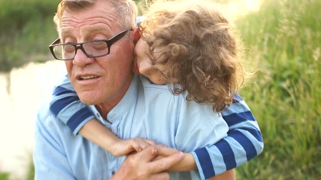 Grandfather and grandson rest, sitting on the river bank. A man tells his curly grandson story. The boy listens attentively. Both laugh joyfully