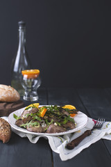 A delicious healthy dinner, a warm chicken liver salad with oranges and rucola, on a black wooden background. A bottle of white wine and fresh bread with avocado. Free space for text. Copy sapace.