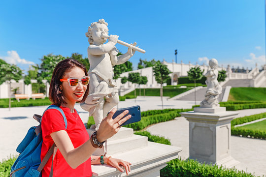 Young female traveler making selfie with smart phone with Cupid statue in summer garden or park