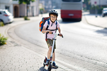 Active school kid boy in safety helmet riding with his scooter in the city with backpack on sunny day. Happy child in colorful clothes biking on way to school.