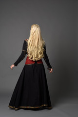 full length portrait of pretty blonde lady wearing  a red and black fantasy medieval gown. standing pose on grey background.