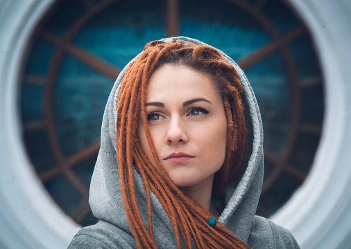 Portrait of a girl with dreadlocks of red color in a white sweatshirt with a hood on the background of a large round window