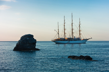 Fototapeta na wymiar Big rock and sailing ship at sunset in front of city harbor, Skopelos island in Greece