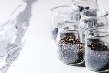 Fototapeta na wymiar Variety of different black peppers allspice, pimento, long pepper, monks pepper, peppercorns and ground powder in glass jars on white marble kitchen table.