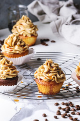Fresh baked homemade cupcakes with coffee buttercream and caramel standing on cooling rack with eucalyptus branch and coffee beans above over white marble kitchen table.