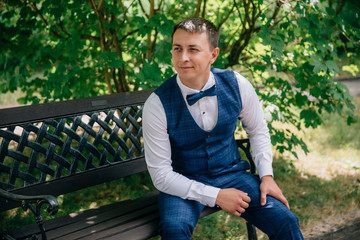 Fototapeta premium A young guy in a stylish fashionable suit with a vest and a bow tie is sitting in the park on a bench. The groom looks away at the bride approaching him and smiles gently, from which he has a dimple