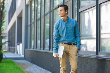 Fototapeta premium handsome smiling young man holding laptop and looking away while standing outside