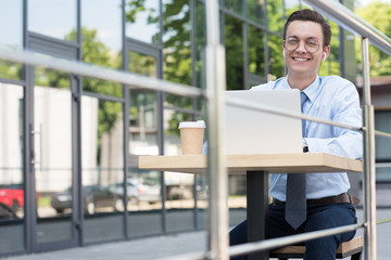 Fototapeta premium handsome smiling young freelancer in eyeglasses using laptop outside modern building