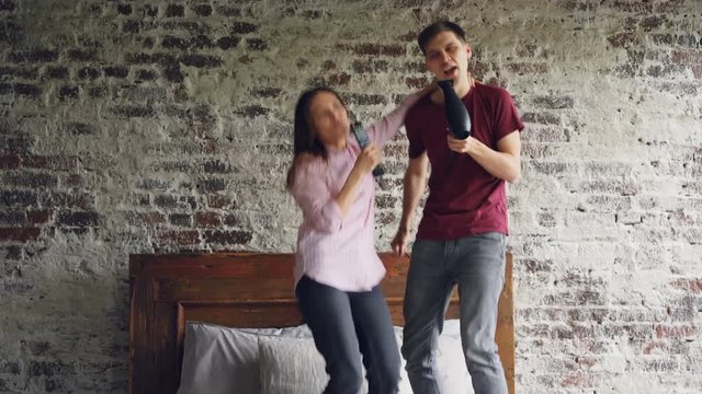 Playful Young People Man And Woman Are Dancing On Bed Listening To Music And Singing Using Hair Dryer And TV Remote As Microphones Having Fun Together.