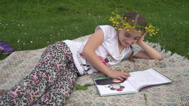 Adorable girl with wreath on the head reading book. Child lying in grass
