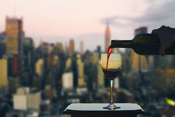 Female hand with bottle pours red wine into glasses on Manhattan background. New York city. ...